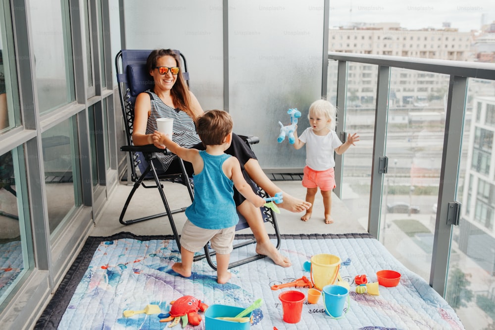 Junge Mutter verbringt Zeit zusammen mit Kindern Babys auf dem Balkon zu Hause. Aufenthalt während der Coronavirus-Covid-19-Pandemie in der Welt. Vorbeugende Maßnahmen gegen die Ausbreitung von Viren.