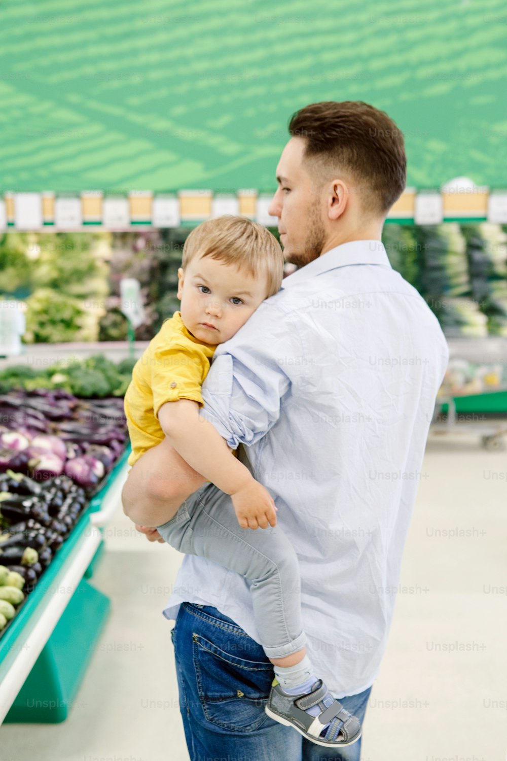 Caucasian father shopping in grocery store with baby son. Dad buying fresh vegetables. Man parent with toddler kid choosing healthy meal for snack lunch. Lifestyle authentic candid moment.