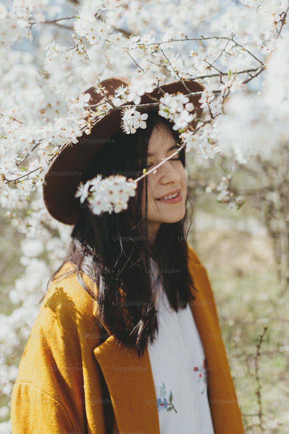 Blooming cherry branches in sunny spring garden on background of stylish beautiful woman in hat sensually posing. Calm tranquil moment of young female embracing in white flowers