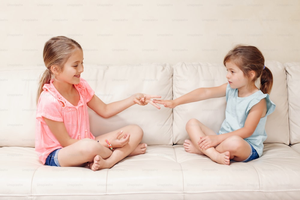 Two girls friends playing rock paper scissors hand game. Caucasian children sitting on couch playing together. Interesting entertaining activity for kids. Authentic candid lifestyle moment.
