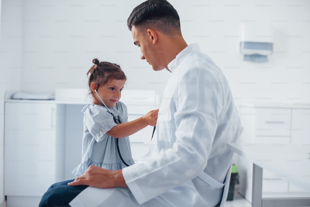 Teaches girl how to use stethoscope. Young pediatrician works with little female visitor in the clinic.