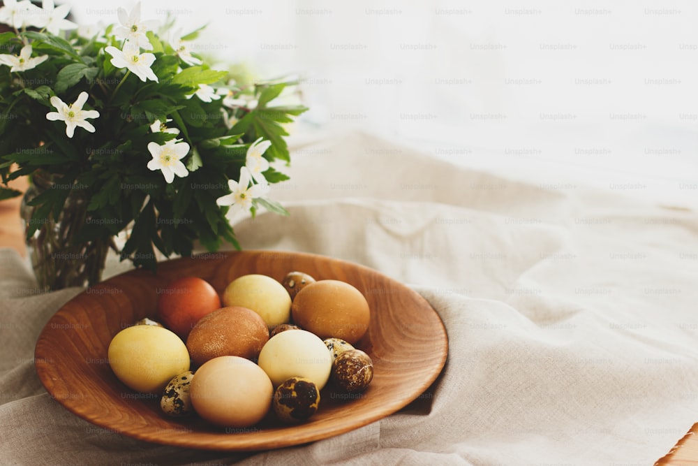 Happy Easter! Modern easter eggs in wooden bowl on rustic linen cloth with spring flowers. Natural dyed eggs in yellow and red colors on rural table with blooming flowers anemone. Aesthetic