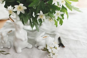 Cute white bunnies and spring flowers on linen cloth napkin on rustic table in soft light. Happy Easter ! Easter hunt concept. White rabbit figurines and blooming anemones flowers rural still life