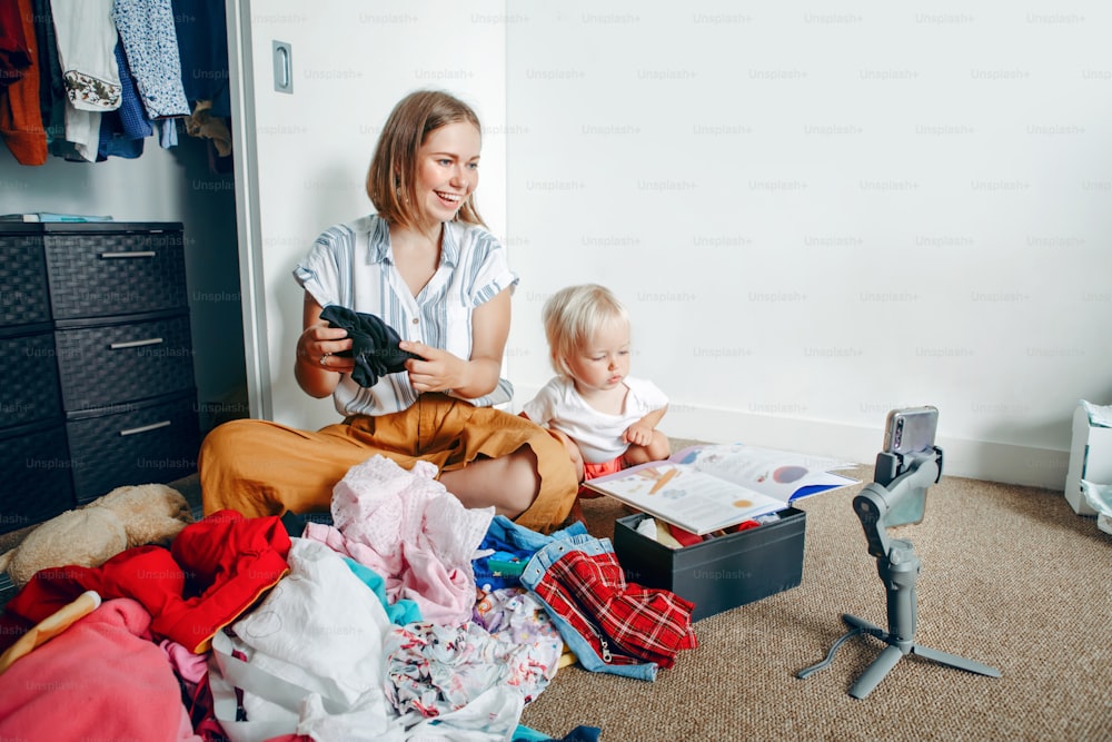 Young mother blogger doing video live stream for social media while sorting clothes at home. Video call chat with friends and family on smartphone. Woman using gimbal tripod for video and phone calls.