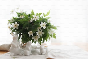 Buona Pasqua! Simpatici coniglietti bianchi e fiori primaverili su tovagliolo di stoffa di lino su tavolo rustico in luce soffusa.  Concetto di caccia pasquale. Figurine di coniglio bianco e anemoni in fiore fiori natura morta rurale