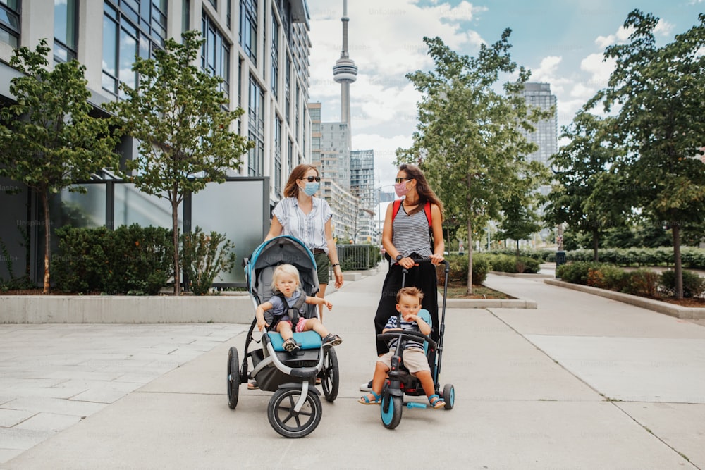 Two Caucasian moms with strollers and kids walking together in Toronto city Canada. Women in face masks with children outdoor. Friends talking on street keeping social distance. New normal.