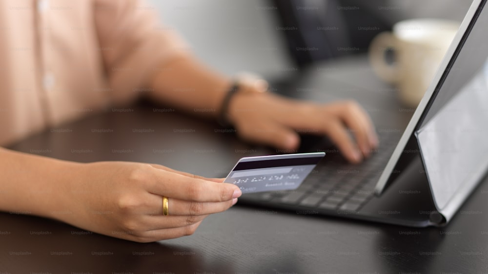 Side view of female holding debit card and using digital tablet for online paying on the table