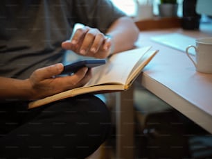 Vista de cerca de la mano masculina usando un teléfono inteligente mientras lee un libro en el escritorio de la computadora en la oficina en casa