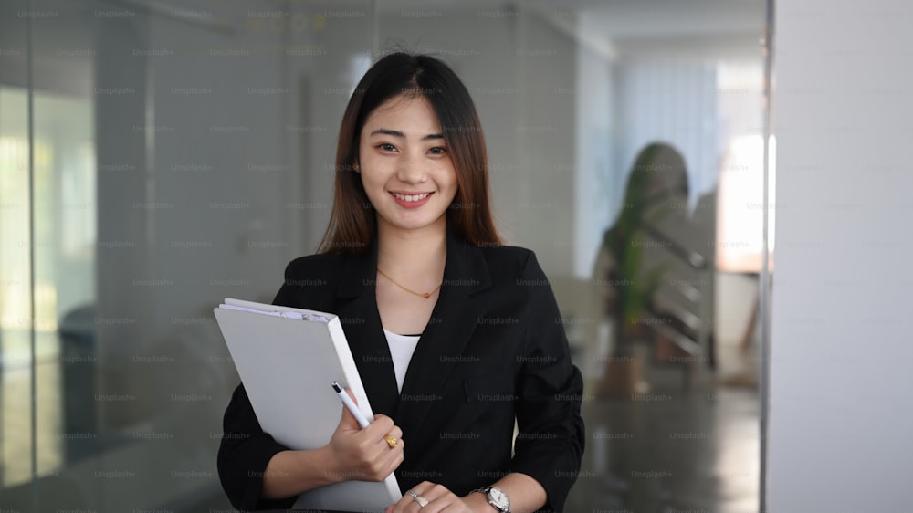 Cheerful young woman office worker holding folder and smiling to camera while standing in office.