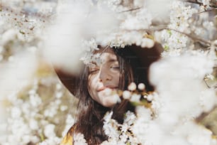 Mujer hermosa con estilo en sombrero que posa sensualmente entre las ramas de cerezo en flor en primavera soleada, momento tranquilo. Retrato de una mujer joven de moda en chaqueta amarilla abrazando en flores blancas