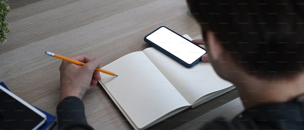 Horizontales Bild eines Mannes, der Notizen auf einem leeren Notizbuch macht und ein Mobiltelefon auf einem Holztisch benutzt.