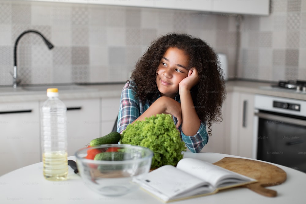 Close up image of cute teen curly mixed-race dreamy girl, cooking salad with fresh vegetables, sitting at the tables in light kitchen, smiling and looking at the window.