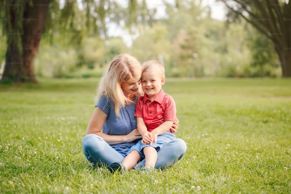 Mothers Day holiday. Young smiling Caucasian mother and laughing boy toddler son sitting on grass in park. Family mom and child hugging having fun together. Happy authentic family lifestyle.