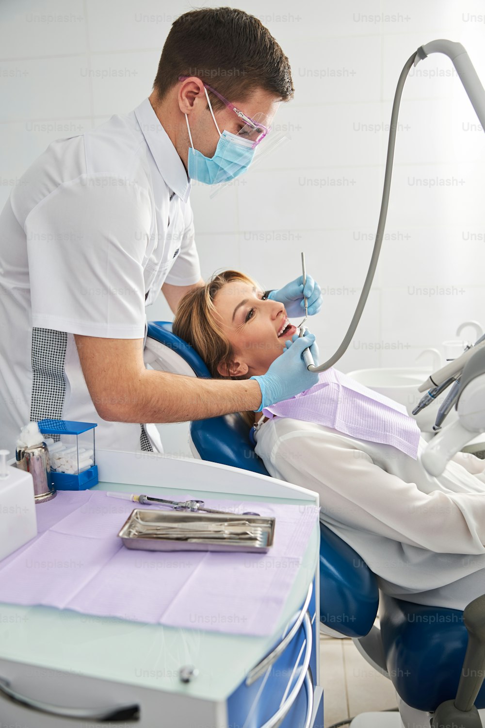 Dentist in a facial mask treating the teeth of a female under operation by using dental tools