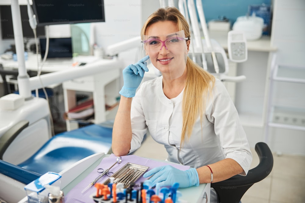 Dental surgeon placing right hand finger to her cheek with another arm lying on a table with dental tools