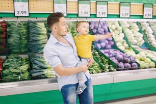 Caucasian father shopping in grocery store with baby son. Dad buying fresh vegetables. Man parent with toddler kid choosing healthy meal for snack lunch. Lifestyle authentic candid moment.