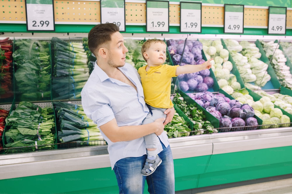 Père caucasien faisant ses courses à l’épicerie avec son fils bébé. Papa achète des légumes frais. Homme parent avec enfant en bas âge choisissant un repas sain pour le déjeuner de la collation. Style de vie authentique moment candide.