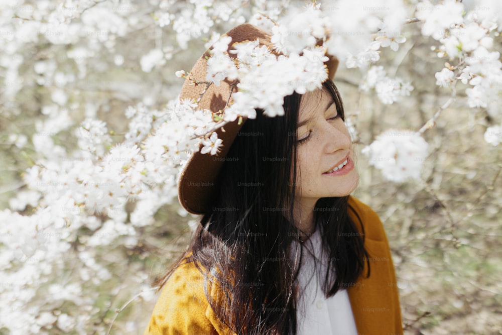 Stylish beautiful woman in hat sensually posing among blooming cherry branches in sunny spring, calm moment. Portrait of happy young brunette female in yellow jacket embracing in white flowers