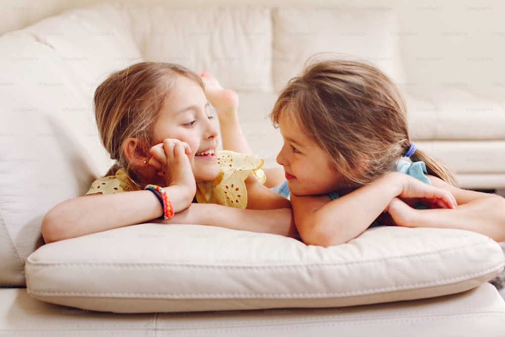 Two cute little Caucasian girls siblings playing at home. Adorable smiling children kids lying on couch together. Authentic candid lifestyle domestic life moment. Happy friends sisters relationship.