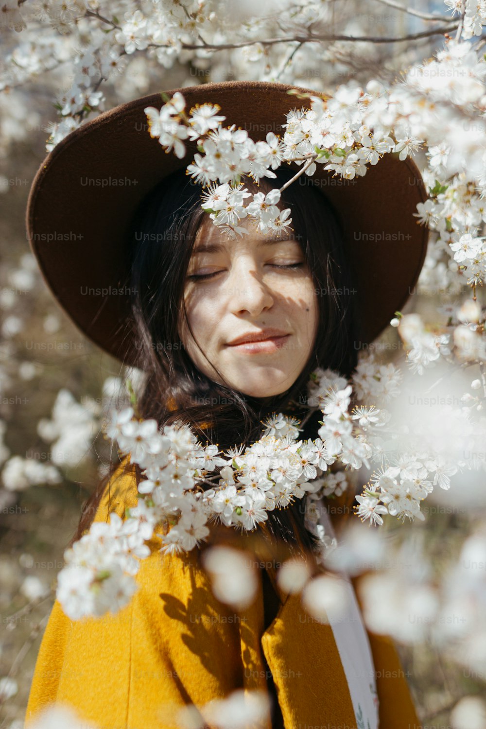 Stylish beautiful woman in hat sensually posing among blooming cherry branches in sunny spring time, calm moment. Portrait of fashionable young female in yellow jacket embracing in white flowers