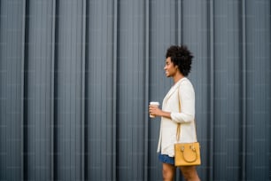 Ritratto di donna d'affari afro che tiene una tazza di caffè mentre cammina all'aperto per strada. Concetto di business e urbano.