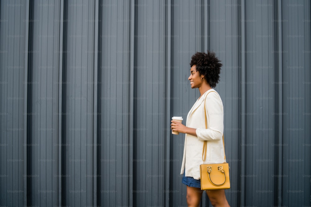 Retrato de una mujer de negocios afro sosteniendo una taza de café mientras camina al aire libre en la calle. Concepto empresarial y urbano.
