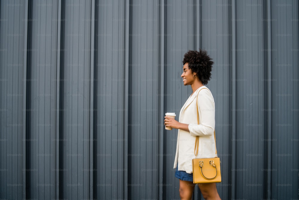 Portrait of afro business woman holding a cup of coffee while walking outdoors on the street. Business and urban concept.