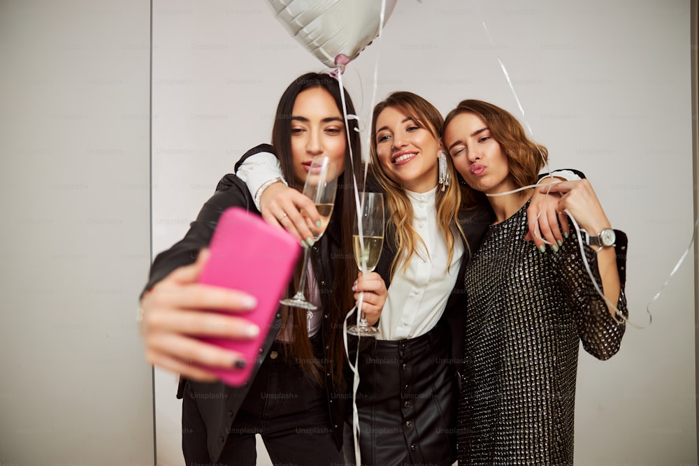 Vue de face d’amies éméchées avec des flûtes à champagne et des ballons posant pour la caméra