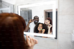 Daily hygiene. Nice happy multiethnic family brushing their teeth while doing morning hygienic procedures. Reflection at the mirror of the family brushing teeth. Stock photo