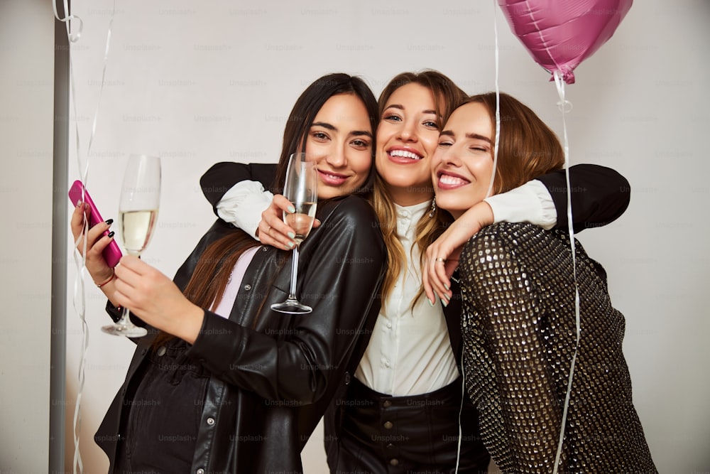 Happy birthday girl with a champagne flute embracing her pleased female friends in front of the camera
