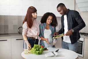 Smiling caucasian mother with her black husband and mixed race daughter cooking salad together, putting chopped fresh vegetables in the glass bowl.