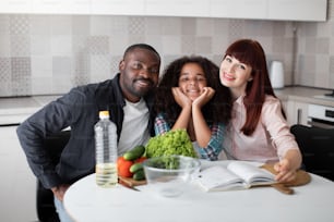 We choosing healthy food. Waist up portrait of the happy multiracial family sitting at the table while looking recipes at the book. Three people looking at the camera with pleasure smiles
