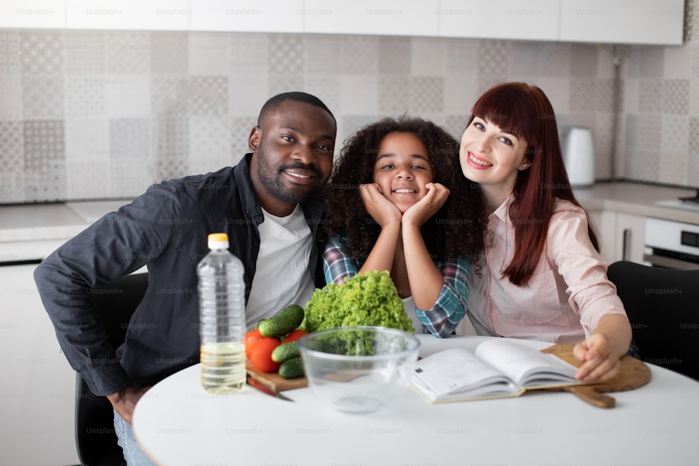 Wir wählen gesunde Lebensmittel. Porträt der glücklichen multiethnischen Familie, die am Tisch sitzt und Rezepte auf das Buch schaut. Drei Menschen, die mit einem Lächeln in die Kamera schauen