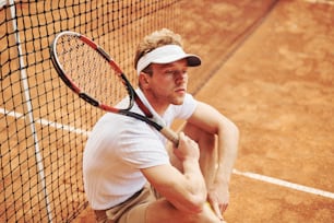 Tired tennis player in sportive clothes is on the court outdoors leaning on the net.