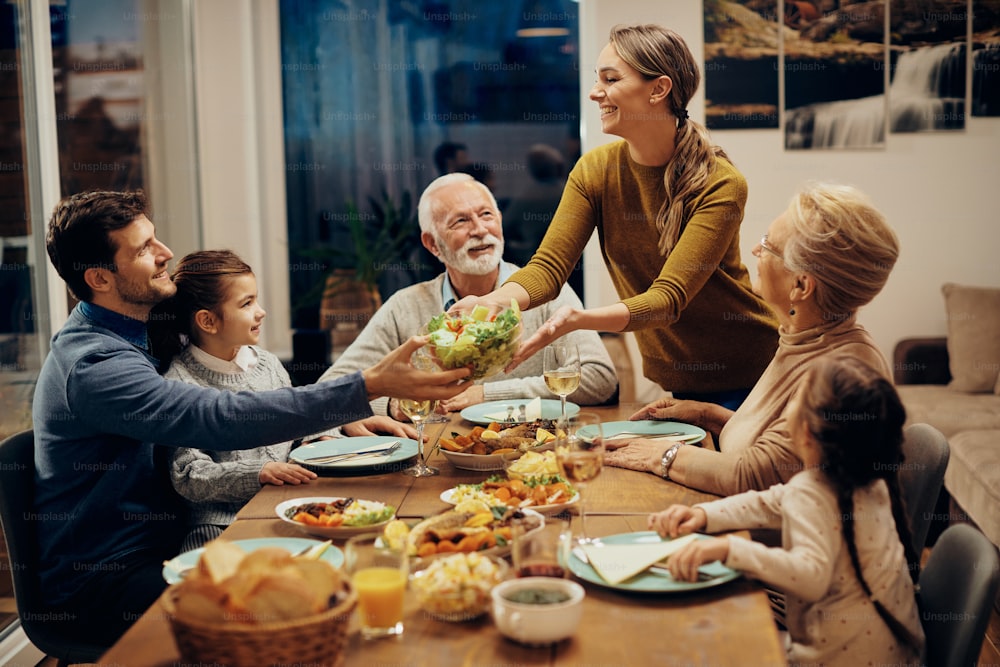 Feliz familia multigeneracional disfrutando de un almuerzo juntos en casa. El foco está en la mujer joven que sirve ensalada en la mesa del comedor.