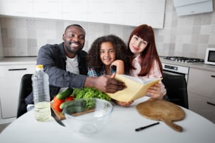 Book about healthy food. Happy adorable multiracial family of three looking at the camera with pleasure smiles while preparing fresh salad at the kitchen. Stock photo