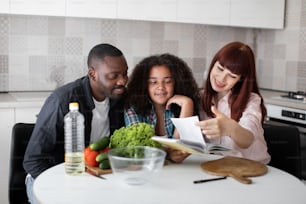 Loving Caucasian mother and African father teaching teenage daughter cooking healthy food at home kitchen. Caring parents and teen girl looking recipes at the book while preparing salad for dinner