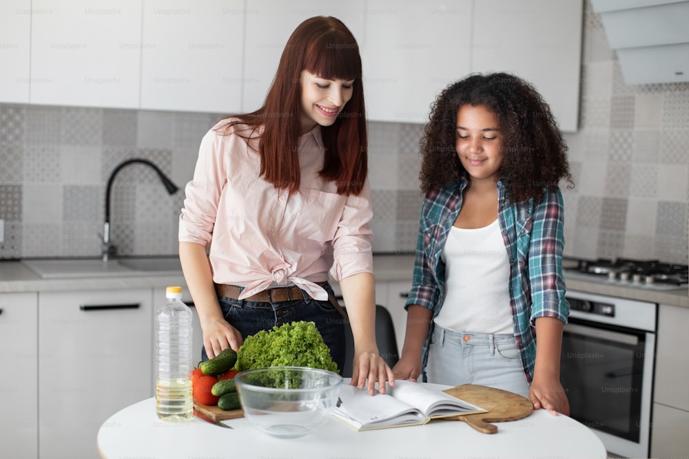 Mixed raced girl daughter preparing lunch with her caucasian mom, while ingredients for salad laying on table. Healthy lifestyle, teach kid cook, motherhood concept