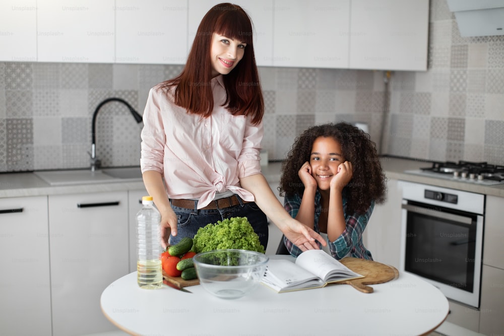 Multiracial daughter preparing lunch with her caucasian mother while looking at the recipes at the book. Ingredient for salad on table. Healthy lifestyle, teach kid cook, motherhood concept