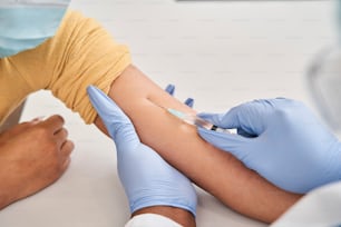 I do not like this. Doctor in sterile gloves giving shot to her female patient wearing protective mask while she sitting at the table. Coronavirus pandemic concept. Stock photo