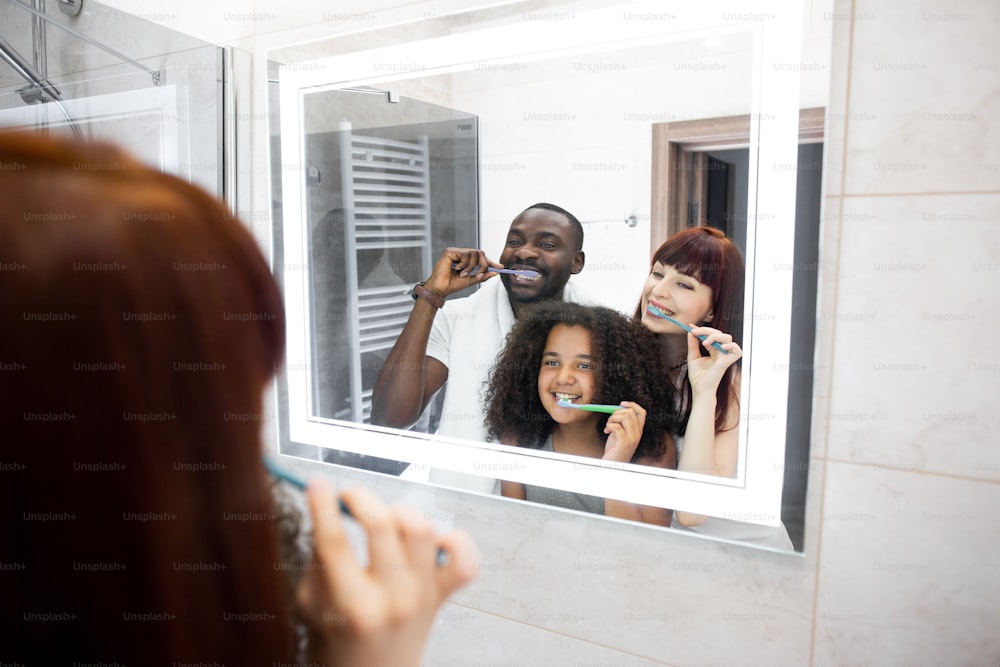 Reflet miroir de la jeune famille multiraciale s’amusant tout en se nettoyant les dents ensemble dans une salle de bain moderne. Des conjoints positifs souriants et joyeux et leur fille faisant une routine d’hygiène bucco-dentaire matinale