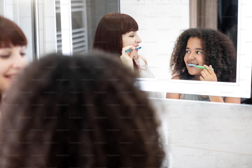 Reflexo espelhado da jovem mãe caucasiana e sua filha adolescente mestiça se divertindo enquanto limpam os dentes juntos no banheiro moderno.
