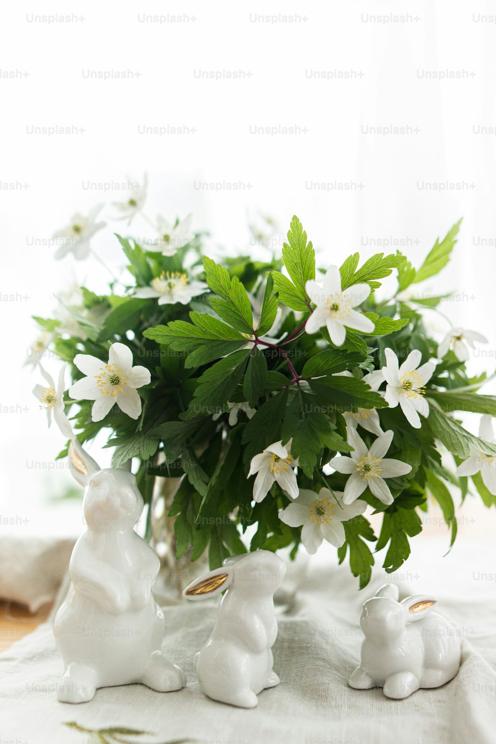 Cute white bunnies and spring flowers on linen cloth napkin on rustic table in soft light. Happy Easter ! Easter hunt concept. White rabbit figurines and blooming anemones flowers rural still life