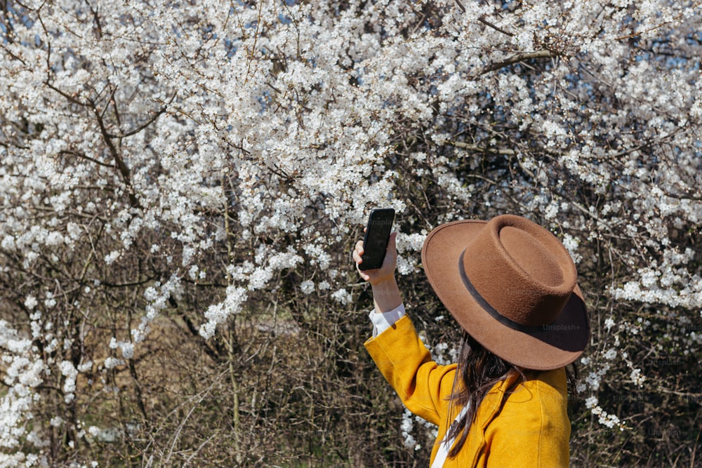 Stylish beautiful woman in hat holding phone at blooming cherry branches in sunny spring. Fashionable young female in yellow jacket taking photo of white flowers or chatting online