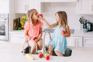 Kaukasische Kindermädchen, die frisches Obst essen, sitzen in der Küchenspüle. Glückliche Familienschwestern Geschwister beim Snack. Bio-Lebensmittel gesunde, leckere leckere Mahlzeit für Kinder. Lifestyle authentischer Moment