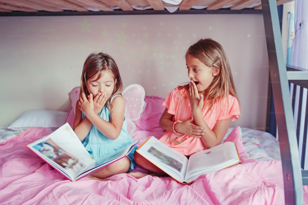 Sorprendidas y asustadas hermanas caucásicas leyendo libros en el dormitorio. Los niños que educan en casa a los hermanos aprendieron que veían lo inesperado en el libro. Educación, desarrollo, aprendizaje. Estilo de vida auténtico de la infancia.
