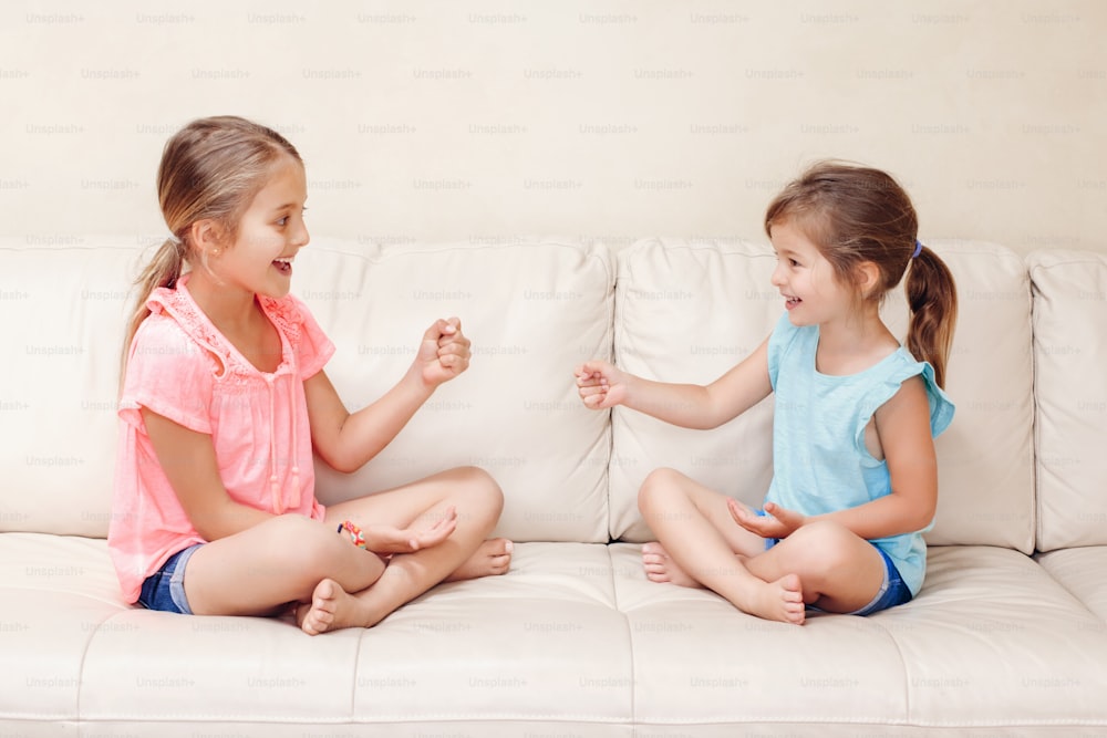 Zwei Freundinnen spielen Steinpapierscheren-Handspiel. Kaukasische Kinder sitzen auf der Couch und spielen zusammen. Interessante unterhaltsame Aktivität für Kinder. Authentischer offener Lifestyle-Moment.