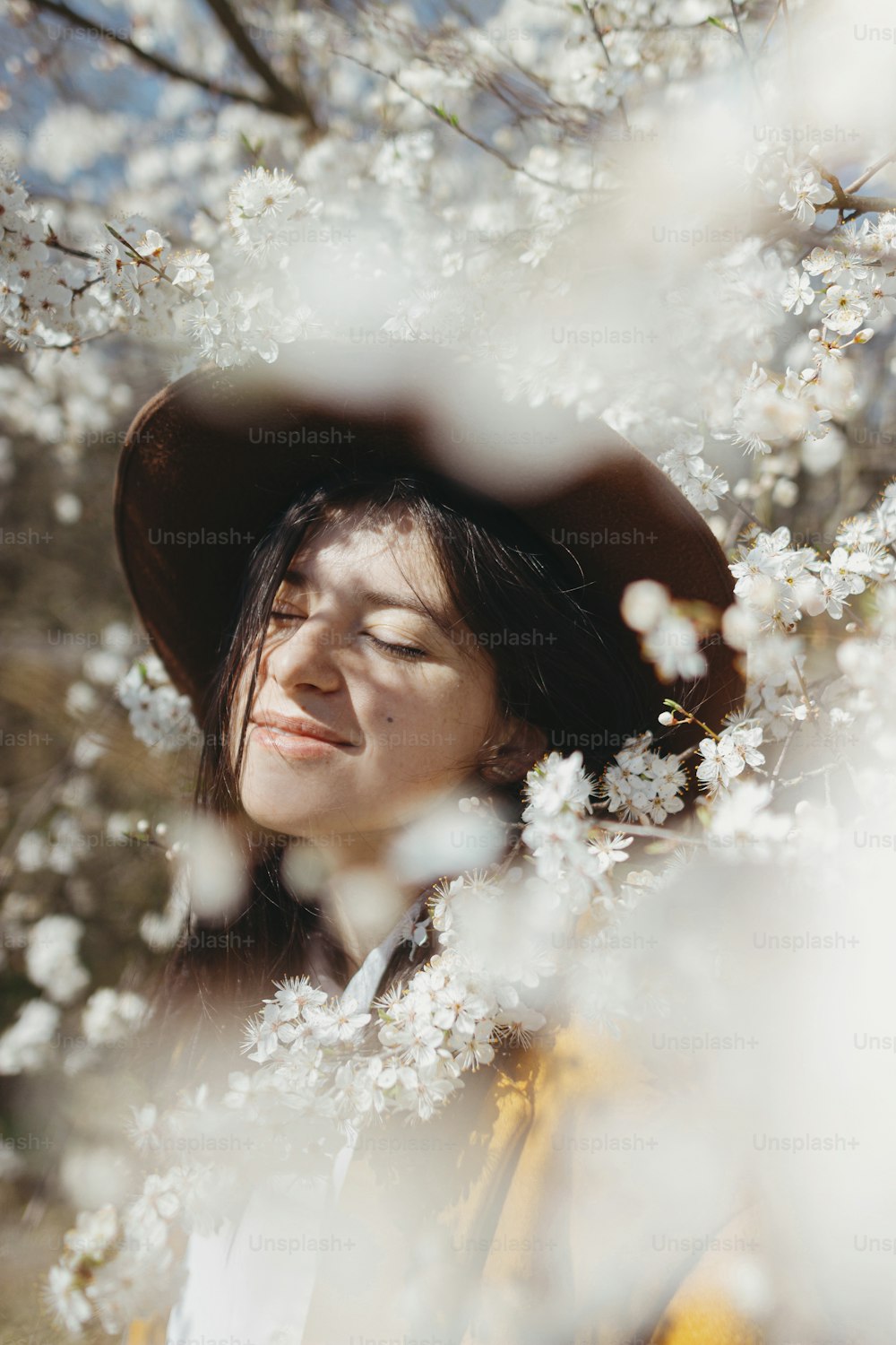 Portrait of happy young brunette female in yellow jacket embracing in white flowers.Stylish beautiful woman in hat sensually posing among blooming cherry branches in sunny spring, calm moment