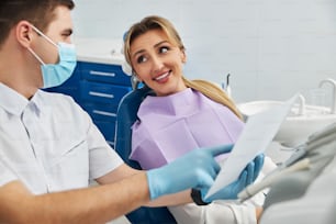 Femme aux cheveux longs allongée sur une chaise de dentiste et regardant avec espoir un médecin, qui montre ses dents radiographie