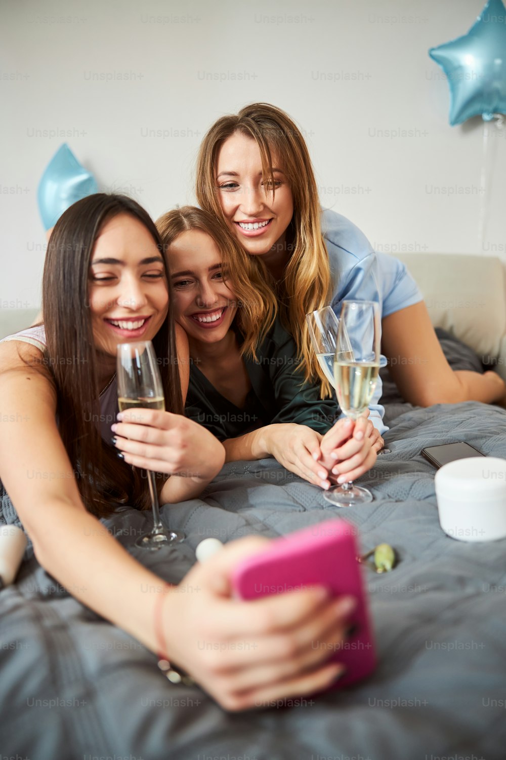 Companhia de três mulheres caucasianas alegres com taças de vinho espumante tirando selfies na cama